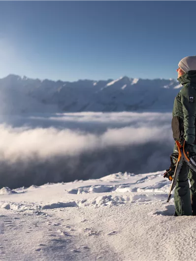 Eine Person steht auf einem schneebedeckten Gipfel und schaut über die Wolken zu den Bergen. Die Szene ist hell erleuchtet und vermittelt ein Gefühl von Freiheit und Abenteuer.