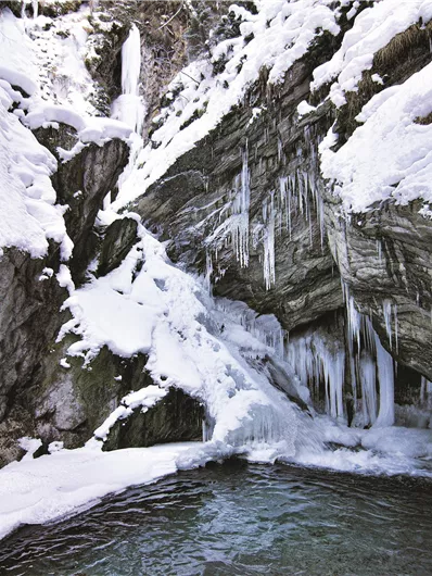 Ein gefrorener Wasserfall umgeben von Schnee und gefrorenen Felsen. Im Vordergrund ist ein klarer Wasserpool sichtbar.