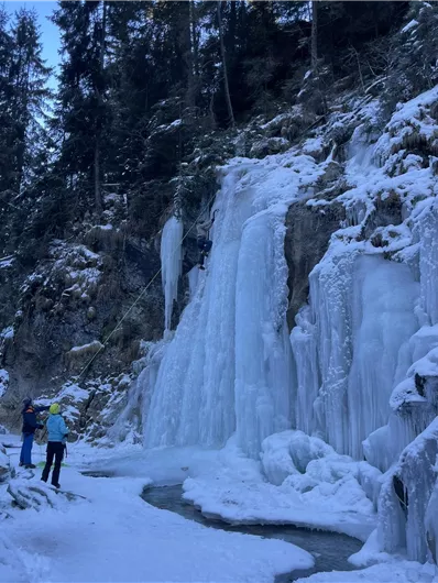 A frozen waterfall with ice cascades in a wintry landscape. Two people are watching a climber ascending the ice wall.
