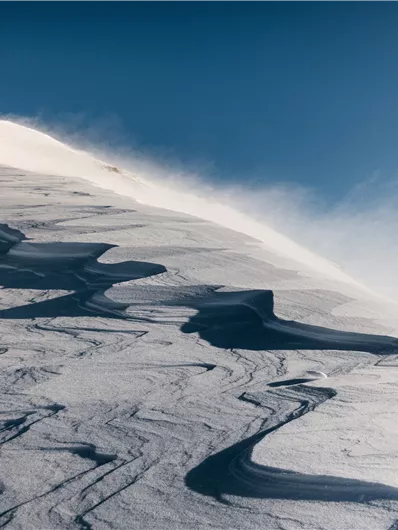 A snowy mountain landscape with gentle waves and a clear blue sky. The wind swirls the snow at the surface.