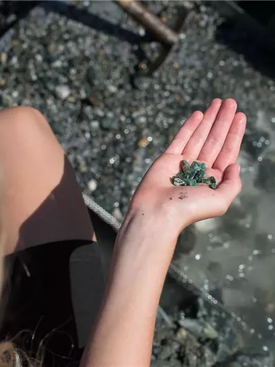 A person is holding some small green stones in their hand. In the background, gravel and water are visible.