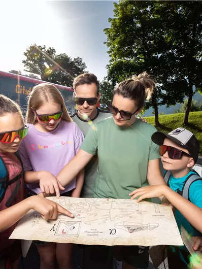 A group of children and adults is together looking at a map. They seem to be preparing for an adventure or a trip.