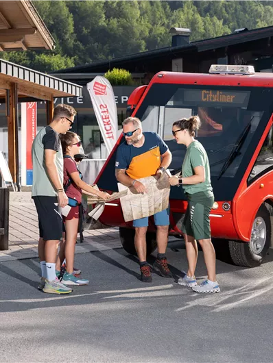 A group of four people stands at a bus stop next to a red shuttle bus. In the background, there is a wooden cabin and green mountains.
