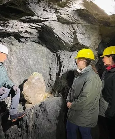 Eine Gruppe von Menschen in einem Bergwerk trägt Helme und schaut auf einen großen Gesteinsbrocken. Der Raum ist dunkel, mit Felsen und einem Licht von oben.