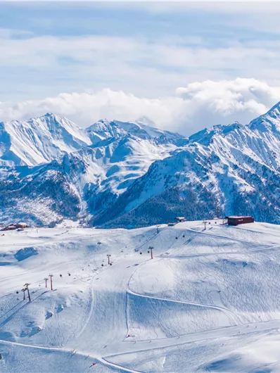 A snow-covered mountain landscape with high peaks and a clear sky. In the foreground, ski lifts and ski slopes can be seen.