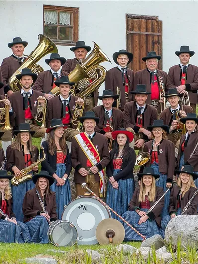 A brass band in traditional attire is standing outdoors. It consists of men and women holding various musical instruments.