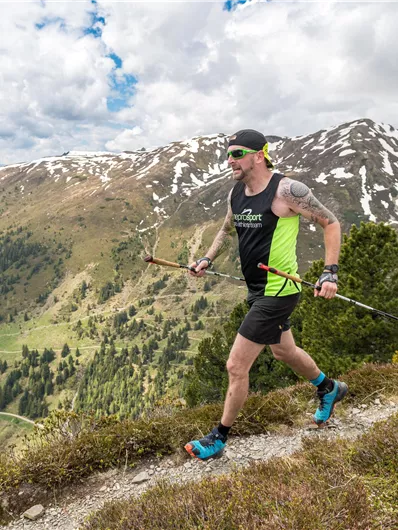 A runner with a pole is walking a hiking trail in the mountains. In the background, snow-capped peaks and green meadows are visible.