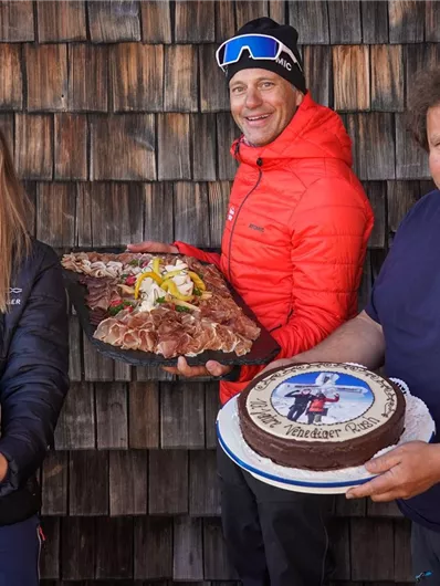 Drei Personen posieren vor einer Holzverkleidung und halten verschiedene Kuchen und eine Fleischplatte in den Händen. Die Atmosphäre ist freundlich und festlich.