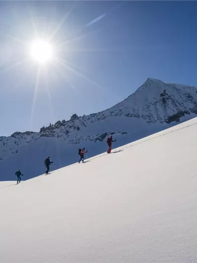 Eine Gruppe von Personen wandert auf einem schneebedeckten Hang. Im Hintergrund ist ein bergiger Gipfel unter klarem blauen Himmel zu sehen.