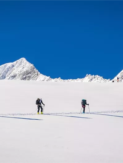 Eine Gruppe von Wanderern bewegt sich über eine schneebedeckte Fläche. Im Hintergrund sind majestätische Berggipfel und ein klarer blauer Himmel zu sehen.