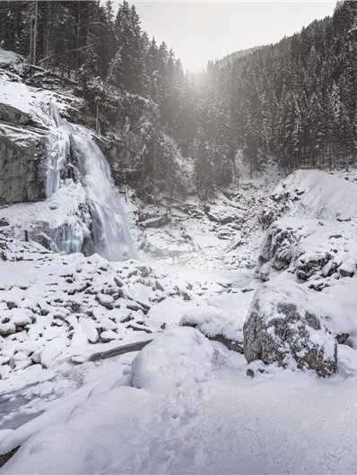 Ein gefrorener Wasserfall umgeben von schneebedeckten Felsen und Bäumen. Die winterliche Landschaft strahlt Ruhe und Kälte aus.