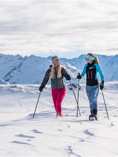 Two women are hiking in the snow with snowshoes. In the background, there are snow-covered mountains and a bright, cloudy sky.