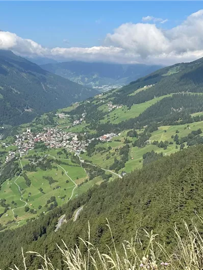 Eine malerische Berglandschaft mit grünen Wiesen und einem kleinen Dorf im Tal. Die Berge sind von Wolken umhüllt und der Himmel ist blau.