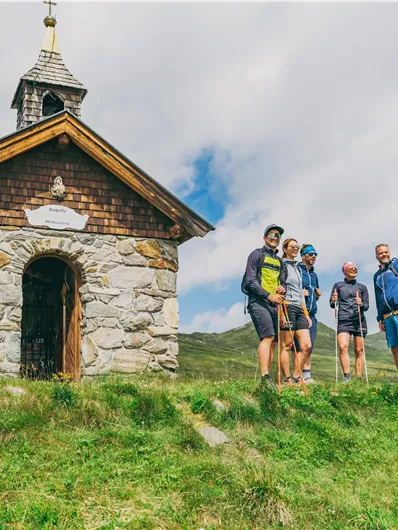 Eine kleine Kapelle steht auf einem Hügel, umgeben von grünen Wiesen. Fünf Wanderer posieren vor der Kapelle und genießen die Aussicht.