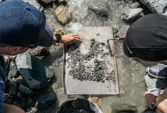 Twee personen zijn bezig om in het water naar gesteente te zoeken. Ze gebruiken een zeef om kleine stenen en mineralen te controleren.
