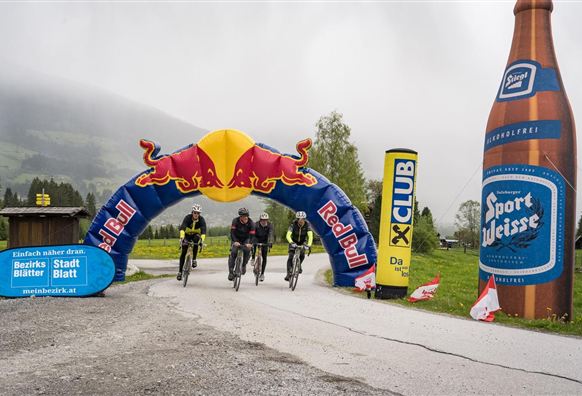 Drei Radfahrer fahren unter einem Roten-Bull-Bogen hindurch. Im Hintergrund sind Nebel und eine große Bierflasche zu sehen.