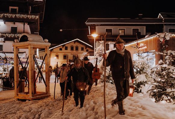 Eine winterliche Szene mit Menschen, die in warmen Kleidern durch den Schnee gehen. Die Umgebung ist festlich beleuchtet und es gibt historische Gebäude im Hintergrund.
