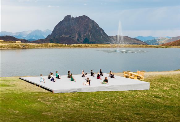 Eine Gruppe von Menschen praktiziert Yoga auf einer Plattform am Ufer eines Sees. Im Hintergrund sind Berge und ein Springbrunnen zu sehen.