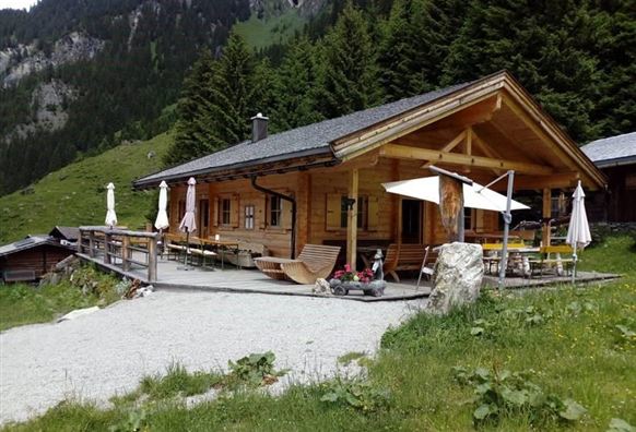 A cozy mountain cabin surrounded by trees and mountains. Outside, there are tables and chairs for guests.