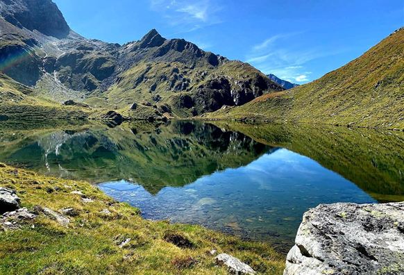 Ein malerischer Bergsee, umgeben von grünen Wiesen und majestätischen Bergen. Der klare Himmel spiegelt sich perfekt im ruhigen Wasser.