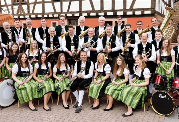 A brass band stands in traditional clothing on a paved area. The musicians are holding various instruments in their hands and smiling at the camera.