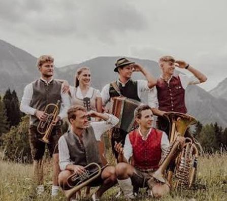 Eine Gruppe junger Musiker in traditioneller Tracht steht im Freien. Sie halten verschiedene Blasinstrumente und genießen die natürliche Umgebung mit Bergen im Hintergrund.