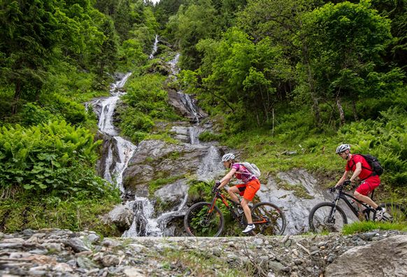 Zwei Mountainbiker fahren entlang eines felsigen Weges, umgeben von üppigem Grün und einem Wasserfall im Hintergrund. Die Szenerie zeigt eine idyllische natürliche Landschaft in den Bergen.
