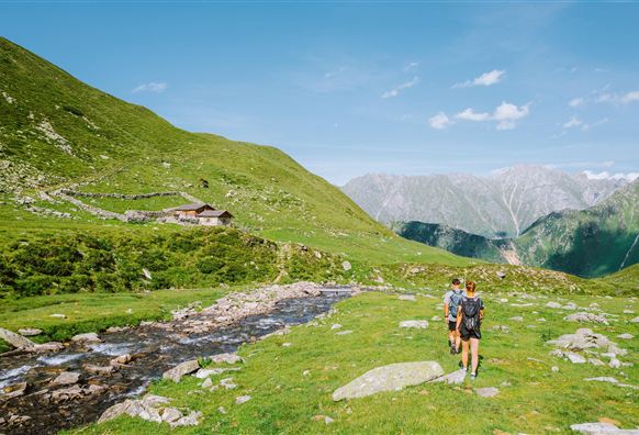 A green valley with a small cabin in the background. Two hikers walk along a clear stream with mountains in the background.