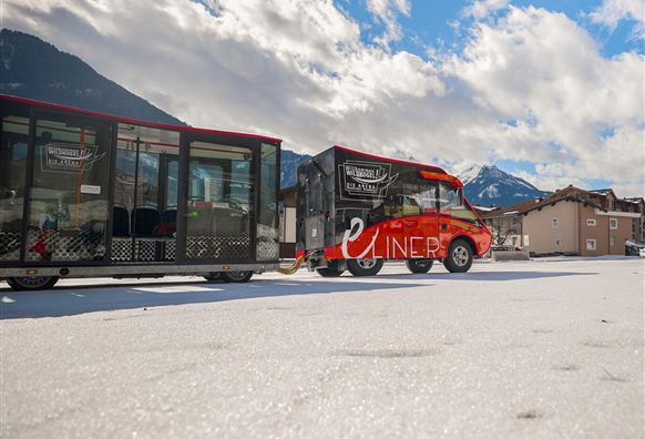 A red electric vehicle with a trailer is standing on a snowy surface. In the background, mountains and a blue sky with clouds can be seen.