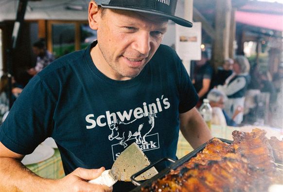 Ein Mann grillt Fleisch und trägt ein T-Shirt mit der Aufschrift "Schweini's". Im Hintergrund sind Menschen und eine große Markise zu sehen.