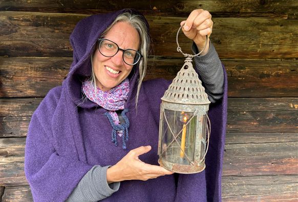 Eine Person in einem lila Umhang hält eine alte Laterne. Im Hintergrund sind Holzwände zu sehen.