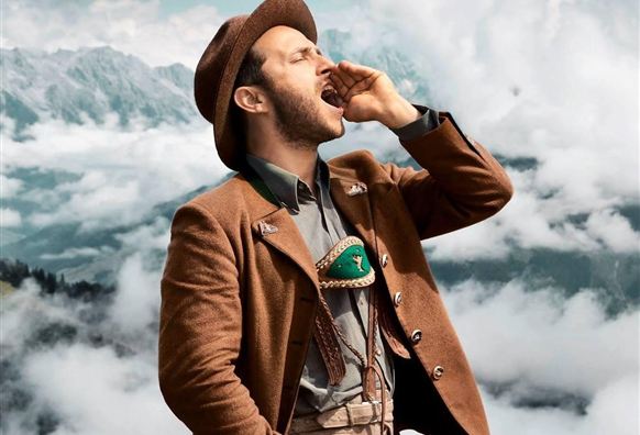 A man in traditional attire is calling out into the landscape. In the background, mountains and clouds can be seen.