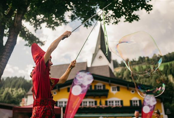 Ein Clown in roter Kleidung zaubert mit riesigen Seifenblasen. Im Hintergrund ist ein buntes Gebäude und Bäume zu sehen.
