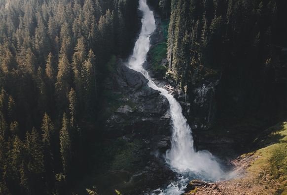 Ein majestätischer Wasserfall fließt über schroffe Felsen inmitten eines dichten Waldes. Umgeben von grünen Wiesen und Bäumen unter einem klaren blauen Himmel.