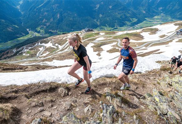Two people are hiking up a steep mountain slope, surrounded by snowy terrain and a picturesque landscape. In the background, green valleys and winding roads can be seen.