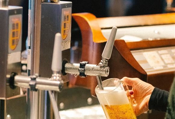 Ein Barmitarbeiter zapft ein Glas Bier an einem Zapfhahn. Im Hintergrund sieht man weitere Gläser und die Bar.