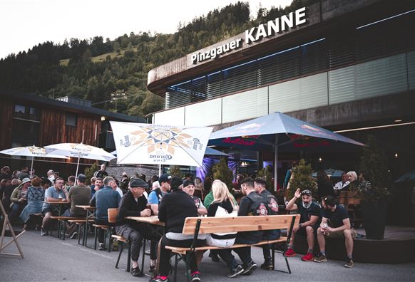 Eine belebte Terrasse vor einem modernen Restaurant mit dem Namen "Pinzgauer Kanne". Menschen sitzen zusammen und genießen Getränke in einer schönen Umgebung.