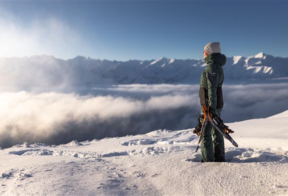 Eine Person steht auf einem schneebedeckten Gipfel und schaut über die Wolken zu den Bergen. Die Szene ist hell erleuchtet und vermittelt ein Gefühl von Freiheit und Abenteuer.
