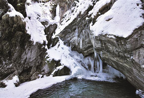 Ein gefrorener Wasserfall umgeben von Schnee und gefrorenen Felsen. Im Vordergrund ist ein klarer Wasserpool sichtbar.
