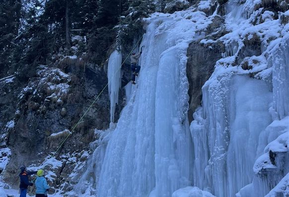 Eine gefrorene Wasserfall mit Eiskaskaden in einer winterlichen Landschaft. Zwei Personen beobachten einen Kletterer, der die Eiswand erklimmt.