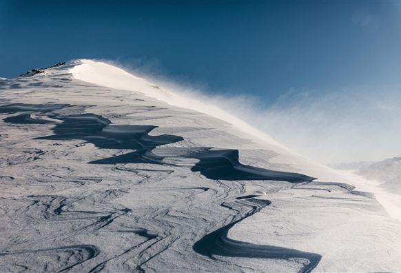 Eine verschneite Berglandschaft mit sanften Wellen und einem klaren blauen Himmel. Der Wind wirbelt den Schnee an der Oberfläche auf.