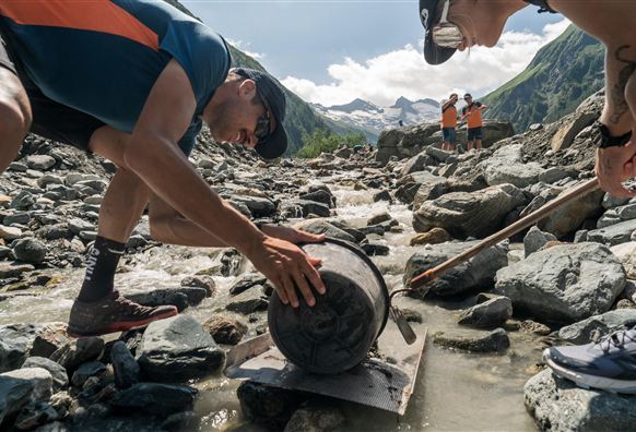 Zwei Personen arbeiten an einem Flussbett und heben einen schweren Gegenstand. Im Hintergrund sind Berge und eine klare Himmelsszene zu sehen.