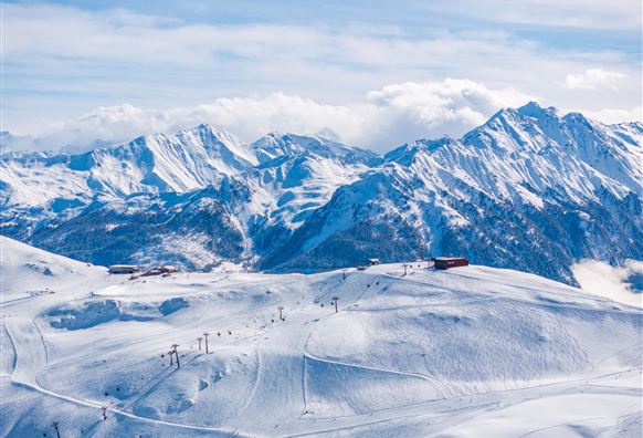 Eine schneebedeckte Berglandschaft mit hohen Gipfeln und einem klaren Himmel. Im Vordergrund sind Skilifte und Skipisten zu sehen.