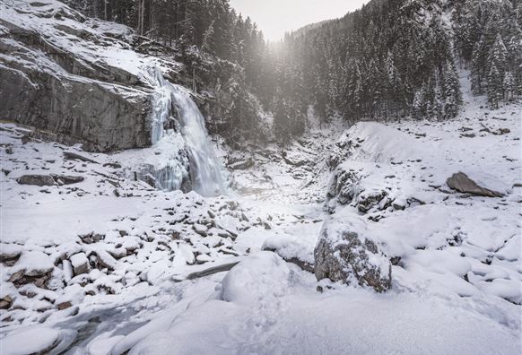 Ein gefrorener Wasserfall umgeben von schneebedeckten Felsen und Bäumen. Die winterliche Landschaft strahlt Ruhe und Kälte aus.