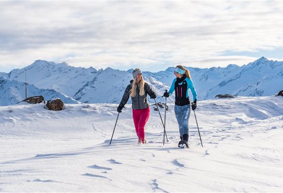 Zwei Frauen wandern im Schnee mit Schneeschuhen. Im Hintergrund sind schneebedeckte Berge und ein heller, bewölkter Himmel zu sehen.