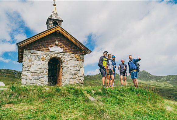 Eine kleine Kapelle steht auf einem Hügel, umgeben von grünen Wiesen. Fünf Wanderer posieren vor der Kapelle und genießen die Aussicht.
