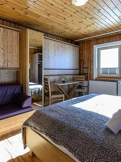 A cozy room with wooden paneling and large windows. There is a bed, a seating area, and a small table.