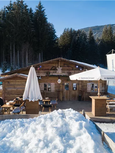 Eine gemütliche Berghütte im Schnee mit Sonnenschirmen und Sitzgelegenheiten. Umgeben von dichten Tannenwäldern unter einem klaren blauen Himmel.