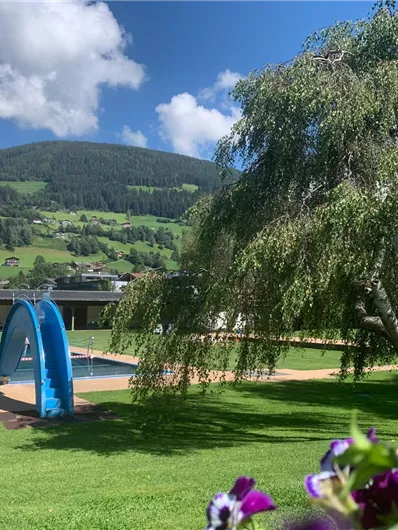 Een mooi buitenzwembad met een glijbaan en groen gras. Op de achtergrond zijn bergen en enkele bomen te zien.