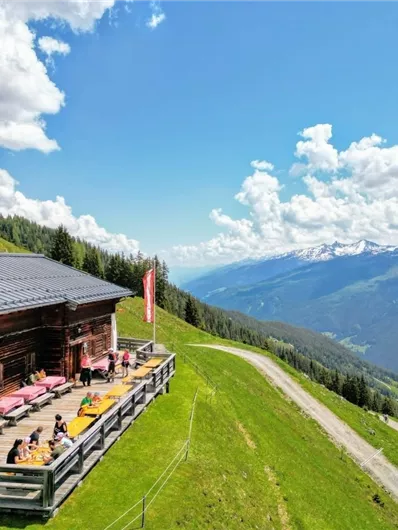 A cozy mountain cabin with a sun terrace on a green meadow. In the background, the mountains stretch under a blue sky with clouds.